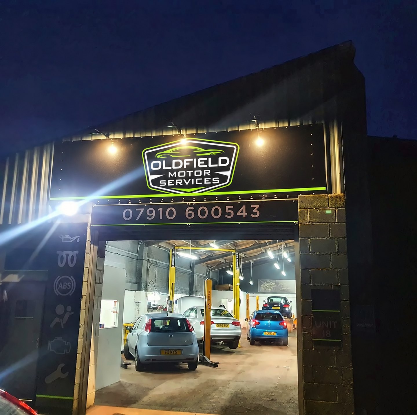 The Oldfield Motor Services workshop at night, sign lit, with cars inside on the ramps
